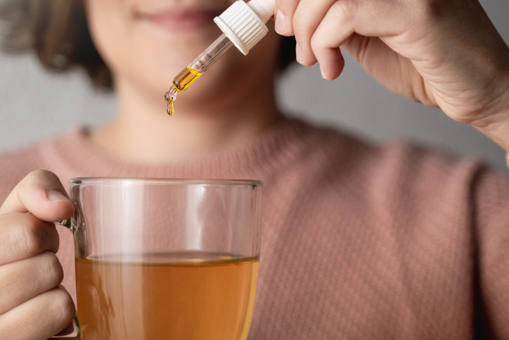 cropped image showing a woman holding a clear mug of tea in one hand and a dropper containing liquid CBD in the other hand, about to add some to the tea